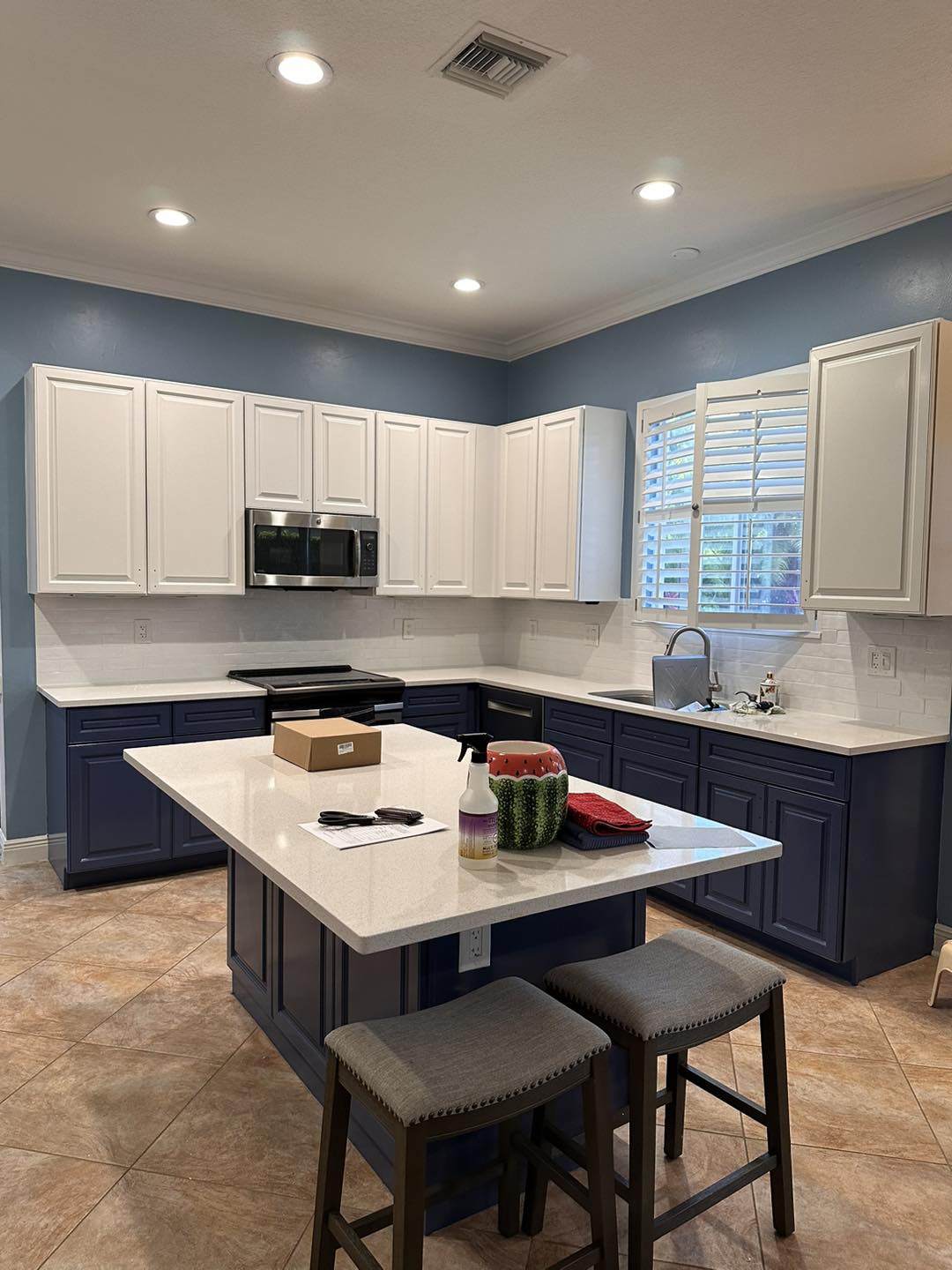 An inviting kitchen boasts a contrasting design with crisp white upper cabinets and elegant navy lower cabinets, finished by KB Painting & Refinishing. The space is enhanced with a white quartz countertop, a classic subway tile backsplash, and warm tile flooring, creating a harmonious blend of classic and contemporary styles in this Port Saint Lucie home.